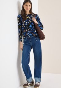 Haut bleu à manches longues avec des motifs de feuilles colorées, associé à un jean ample délavé clair et des baskets marron avec des accents blancs. Sac à main marron porté à l'épaule.