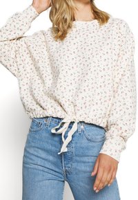 Krémová mikina s květinovým vzorem, volnými rukávy, elastickým lemem a detailem z šňůrky; spárována se světle modrými džíny.