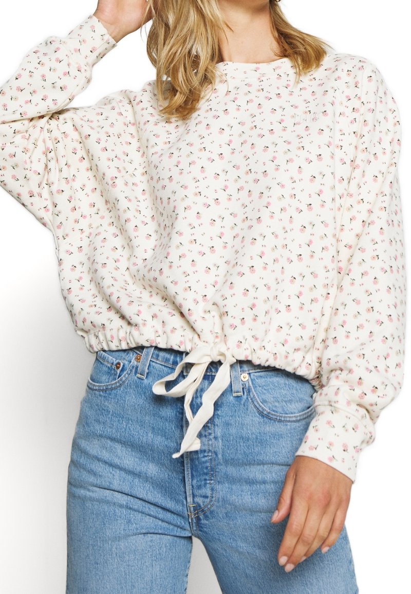 Krémová mikina s květinovým vzorem, volnými rukávy, elastickým lemem a detailem z šňůrky; spárována se světle modrými džíny.