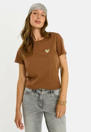 Femme portant un t-shirt marron à manches courtes avec un petit motif cœur, un jean gris clair, un foulard imprimé léopard et des bracelets dorés au poignet droit.