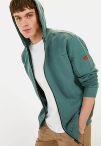 Hoodie zippé vert en tissu doux avec des poignets côtelés, doté d'une poche frontale et d'un patch logo sur la manche gauche. Porté sur un T-shirt blanc.