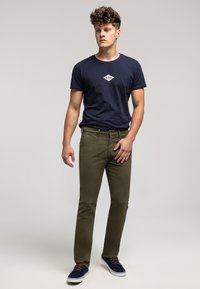 Granatowy bawełniany t-shirt z logo, oliwkowozielone bawełniane spodnie oraz granatowe trampki z brązowymi sznurówkami, stojące na jasnoszarym tle.