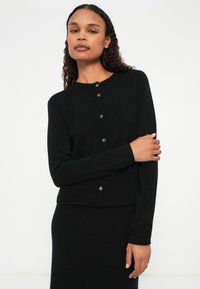 Μαύρο πλεκτό ζακέτα με μακριά μανίκια, στρογγυλή λαιμόκοψη και διακριτική κουμπότρυπα. Μαλακή υφή, κοντή σχεδίαση και εφαρμοστή γραμμή.