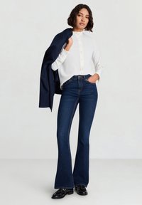 Weiße Bluse mit Rüschenkragen und Knöpfen vorne, kombiniert mit dunkelblauen ausgestellten Jeans. Das Model hält einen marineblauen Blazer und trägt schwarze Schuhe.