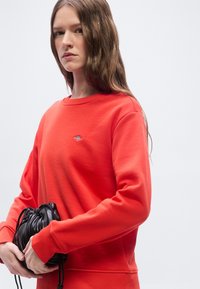 Νεαρή γυναίκα με μακριά καστανά μαλλιά φοράει φωτεινό πορτοκαλί φούτερ και κρατά μικρή μαύρη δερμάτινη τσάντα με πιεστικές λεπτομέρειες, σε απλό γκρι φόντο.