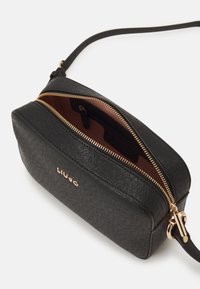 Geantă crossbody din piele neagră cu o suprafață texturată, închidere cu fermoar auriu și un interior maro cu un tag cu logo. Formă compactă rectangulară.