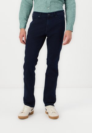 Mørkeblå jeans med slim fit, lavet af denimstof. Har fem lommer og knaplukning. Parret med beige sneakers.