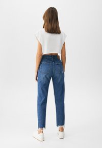 Top blanco recortado, vaqueros azul de talle alto con dobladillos deshilachados y zapatillas blancas, vistos desde atrás. El denim presenta detalles de costuras distintivas.