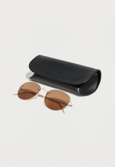 Στρογγυλά γυαλιά ηλίου με καφέ απόχρωση και λεπτό χρυσό σκελετό δίπλα σε κλειστή μαύρη δερμάτινη θήκη Calvin Klein πάνω σε λευκή επιφάνεια.