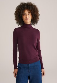 Burgunderfarbener Rollkragenpullover aus weichem Material, taillierte Passform mit langen Ärmeln und geripptem Saum, kombiniert mit blauen weiten Denim-Hosen.