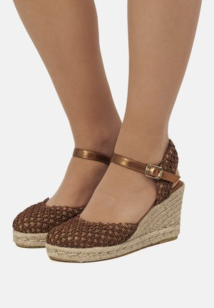 Braune gewebte Keilsandaletten mit Jutesohle und einem metallischen Knöchelriemen mit Schnalle. Rund zulaufende Zehenpartie mit strukturierter Oberseite.
