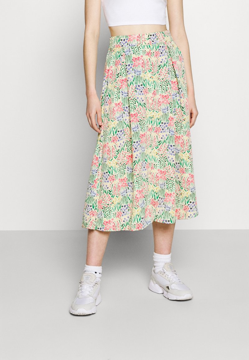 Femme portant une jupe mi-longue fleurie, un crop top blanc, des chaussettes blanches et des baskets blanches, debout devant un mur clair uni.