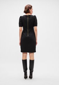 Crna pripijena haljina s kratkim napuhanim rukavima i vidljivim zlatnim patentnim zatvaračem na leđima; nosi se s kožnim čizmama do koljena.