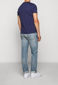 Muž v námořnickém modrém tri čku, světlých džínech s ohrnutými nohavicemi a bílým textem na manžetách, a bílých teniskách, stojící na jednobarevném pozadí.