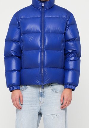 Homme portant une doudoune bleue brillante avec une fermeture éclair à l’avant et un jean bleu clair usé, debout devant un fond uni.