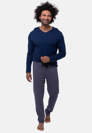 Smilende mand med krøllet hår, der går barfodet, iført en mørkeblå langærmet skjorte og mønstrede pyjamasbukser mod en hvid baggrund.