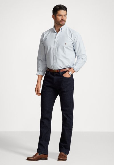 Lys blå og hvid stribet skjorte med knapper, mørkejeans, brun bæltetaske og lædersko. Modellen står med hænderne i lommerne.