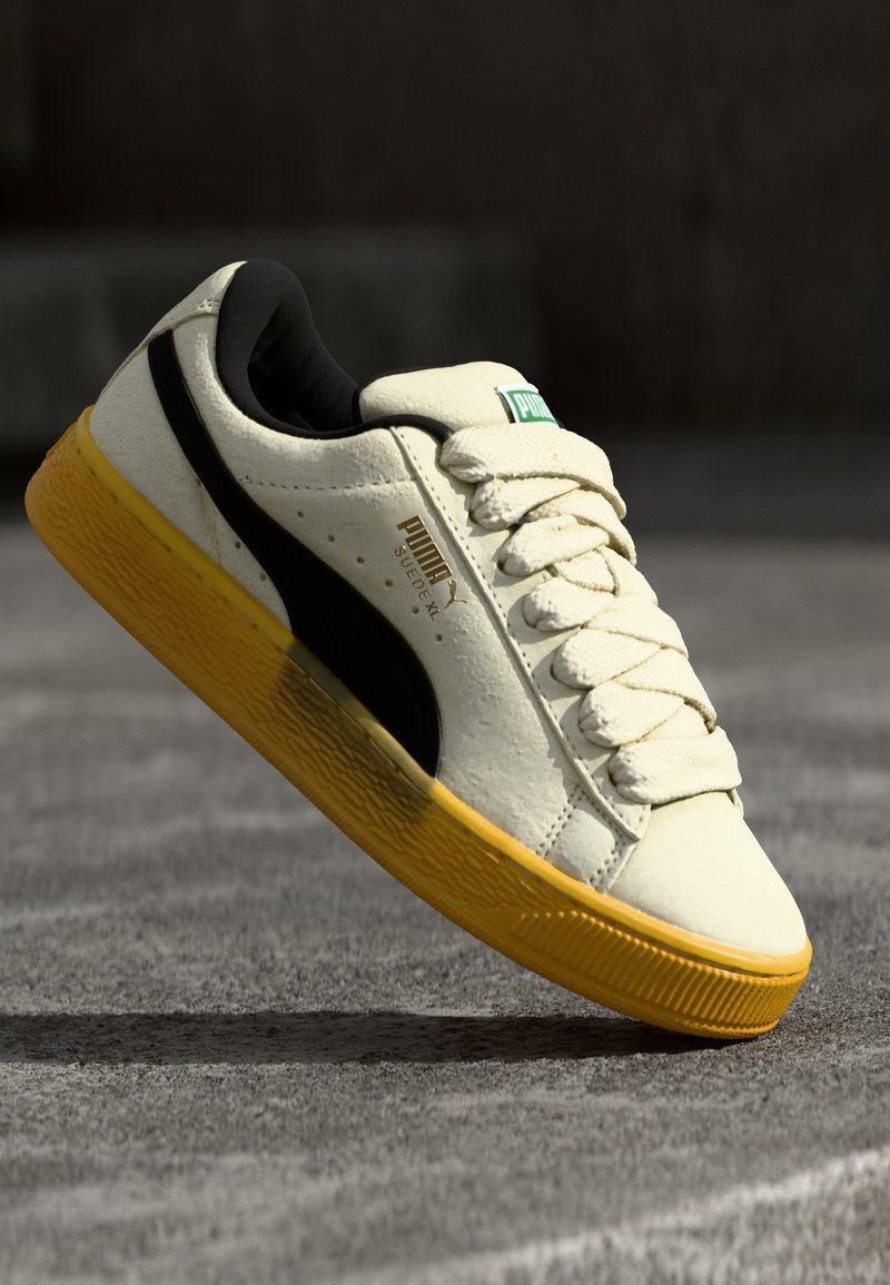Baskets Puma Suede XL blanches avec une bande latérale noire et une semelle jaune, présentées inclinées sur une surface grise.