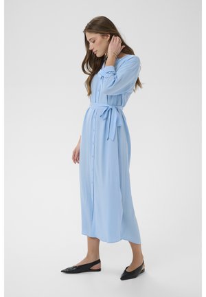 Vrouw met lang bruin haar die een lichtblauwe midi-jurk met knopen draagt, gestrikt in de taille, en zwarte platte slingback schoenen, zijwaarts staand.