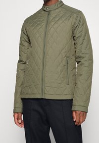 Gepolstertes olivgrünesjack mit Stehkragen, durchgehendem Reißverschluss und Seitentaschen. Verfügt über eine strukturierte Oberfläche und ein elegantes, tailliertes Design.