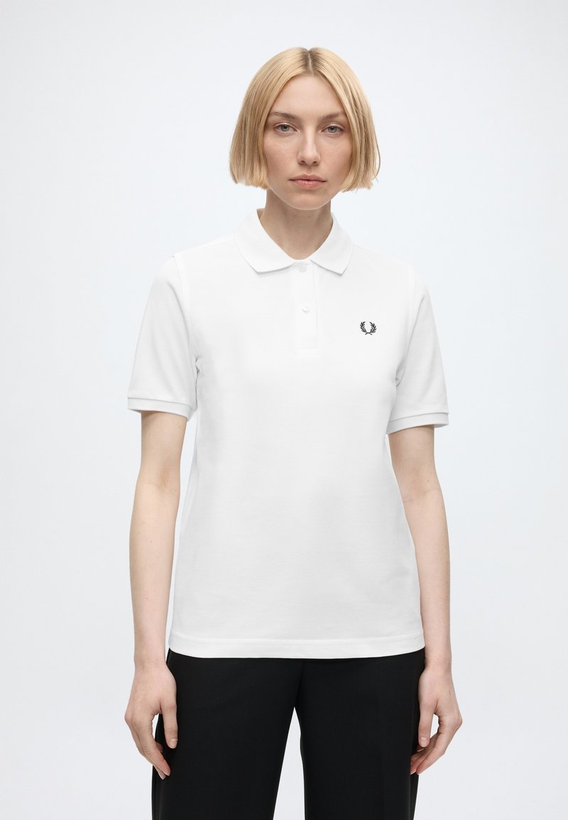 Femme aux cheveux blonds courts portant un polo blanc et un pantalon noir, debout devant un fond clair uni.