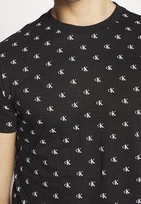 Czarny t-shirt wykonany z bawełny, z powtarzającym się białym wzorem logo 'cK' na całej tkaninie. Krótkie rękawy, standardowy okrągły dekolt.