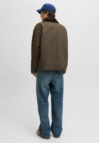 Bruine jas met een donkere kraag, gecombineerd met losse blauwe jeans. Het model draagt een blauwe pet, wat een casual outfitdesign laat zien.