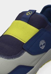 Ναυτικό μπλε, γκρι και κίτρινο αθλητικό παπούτσι με ελαστικό λουρί και λογότυπο Timberland στο πλάι και στη γλώσσα, σε λευκό φόντο.