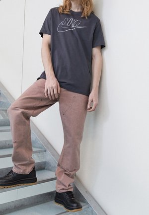 T-shirt Nike gris avec logo blanc, pantalon rose avec coutures visibles et poches, chaussures noires avec semelles texturées, debout sur des escaliers.