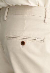 Beige cotton trousers with a back pocket featuring a brown button and a GANT label. The fabric has a subtle texture and clean lines.