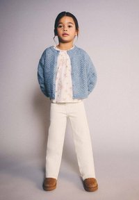 Svetlomodro pleteni jopič preko rožnato vzorčaste svetlo roza majice, kombiniran z belimi hlačami in rjavimi škornji. Preprost dizajn z teksturnimi detajli.