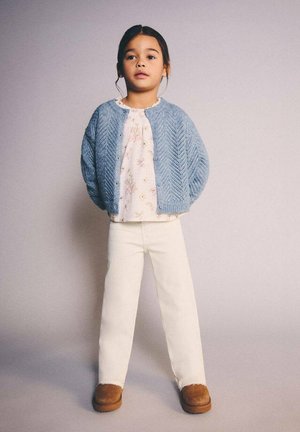 Cardigan azzurro chiaro lavorato a maglia sopra una maglietta rosa pallido con motivo floreale, abbinato a pantaloni bianchi e stivali marroni. Design semplice con dettagli testurizzati.