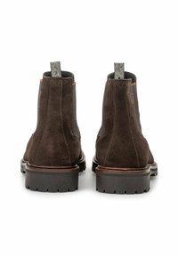 Un paio di stivaletti Chelsea in suede marrone scuro con lati elastici neri, suole spesse con tacche e linguette in tessuto, mostrati da dietro su sfondo bianco.