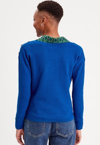 Pull en tricot bleu avec un col à motifs verts, des manches longues et des détails en bouton sur l'épaule. Conçu pour une coupe décontractée.