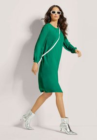 Grünes Kleid mit langen, gefalteten Ärmeln, versehen mit einem weißen Akzentgürtel, kombiniert mit glänzenden, silbernen Ankle Boots mit spitzer Zehe.
