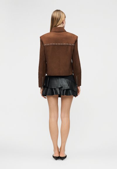 Γυναίκα που στέκεται με την πλάτη προς τα εμπρός, φορώντας καφέ σουέντ μπουφάν με τρουκς, μαύρη δερμάτινη μίνι φούστα με στρώσεις και μαύρα παπούτσια με τακούνι kitten.