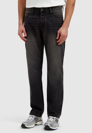 Man die zwarte, vervaagde jeans met rechte pijpen draagt, witte sneakers en een wit overhemd, staand met ontspannen handen langs het lichaam.