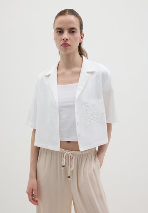 Camiseta blanca recortada con mangas cortas, botones frontales y un bolsillo de encaje texturizado. Llevada sobre una camiseta blanca sin mangas y pantalones beige.