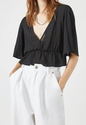 Femme portant un chemisier noir texturé à col en V avec des manches évasées et un pantalon blanc plissé taille haute, les mains dans les poches.