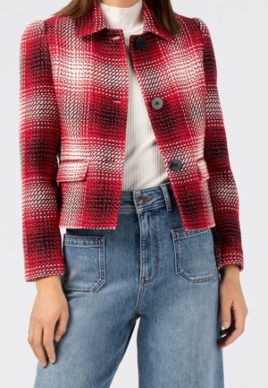 Femme portant une veste à carreaux rouge, noire et blanche boutonnée sur un haut côtelé blanc, accompagnée d'un jean bleu taille haute avec des poches plaquées à l'avant.