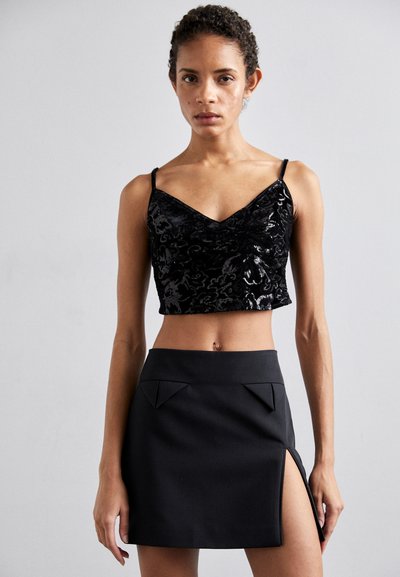 Crop top en velours noir avec un décolleté en V et des fines bretelles, associé à une mini-jupe noire taille haute avec des plis géométriques et une fente sur le côté.