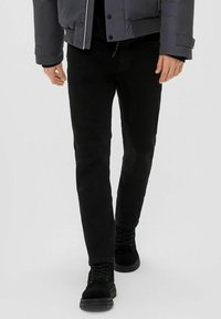 Čierne slim-fit džínsy s hladkou textúrou, doplnené čiernymi šnurovacími čižmami. Oblečenie dopĺňa šedá prešívaná bunda s patentkami.