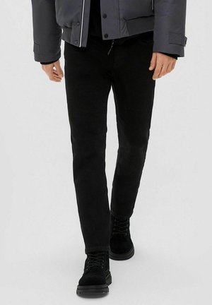 Zwarte slim-fit jeans met een gladde textuur, gecombineerd met zwarte veterschoenen. De outfit bevat een grijze gewatteerde jas met drukknoopsluitingen.