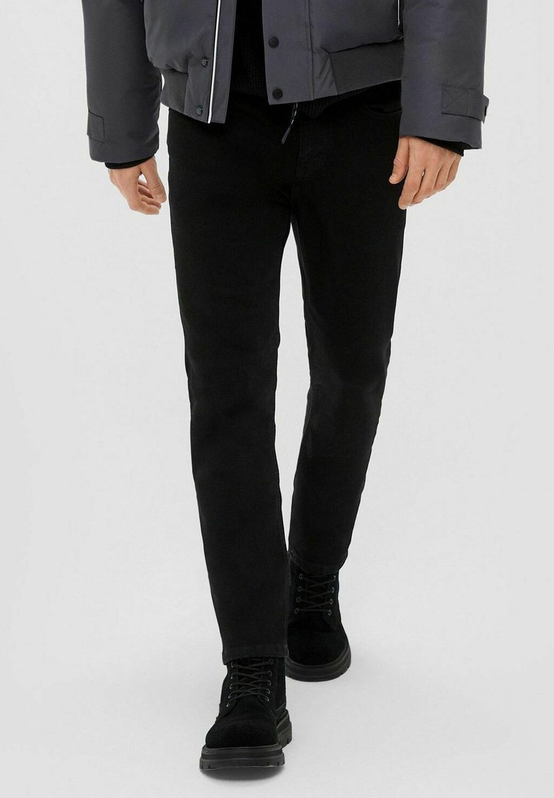 Čierne slim-fit džínsy s hladkou textúrou, doplnené čiernymi šnurovacími čižmami. Oblečenie dopĺňa šedá prešívaná bunda s patentkami.