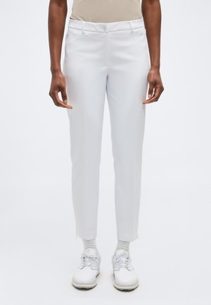 Personne portant un pantalon blanc ajusté avec un bouton à l'avant, des chaussettes blanches et des chaussures blanches à lacets, debout devant un fond uni.