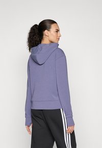 Lila Kapuzenpullover mit langen Ärmeln, Seitenschlitzen und gerippten Bündchen, kombiniert mit schwarzen Hosen mit weißen Streifen. Weiche Textur.
