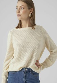Krämfärgad stickad tröja med ett texturerat diamantmönster, vid halsringning och långa ärmar. Matchad med högmidjade blå jeans.