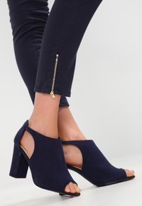 Zapatos de tacón abiertos color azul marino con un diseño recortado, fabricados en un tejido suave, que presentan un tacón grueso y aberturas laterales para mayor transpirabilidad.