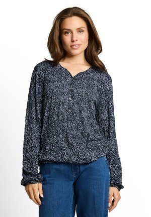 Femme aux cheveux bruns portant un chemisier bleu marine à motifs floraux blancs et un jean bleu, debout devant un fond blanc.