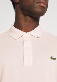 Hombre vistiendo una camiseta polo rosa claro con los botones abrochados, mostrando el logo verde del cocodrilo de Lacoste en el pecho.